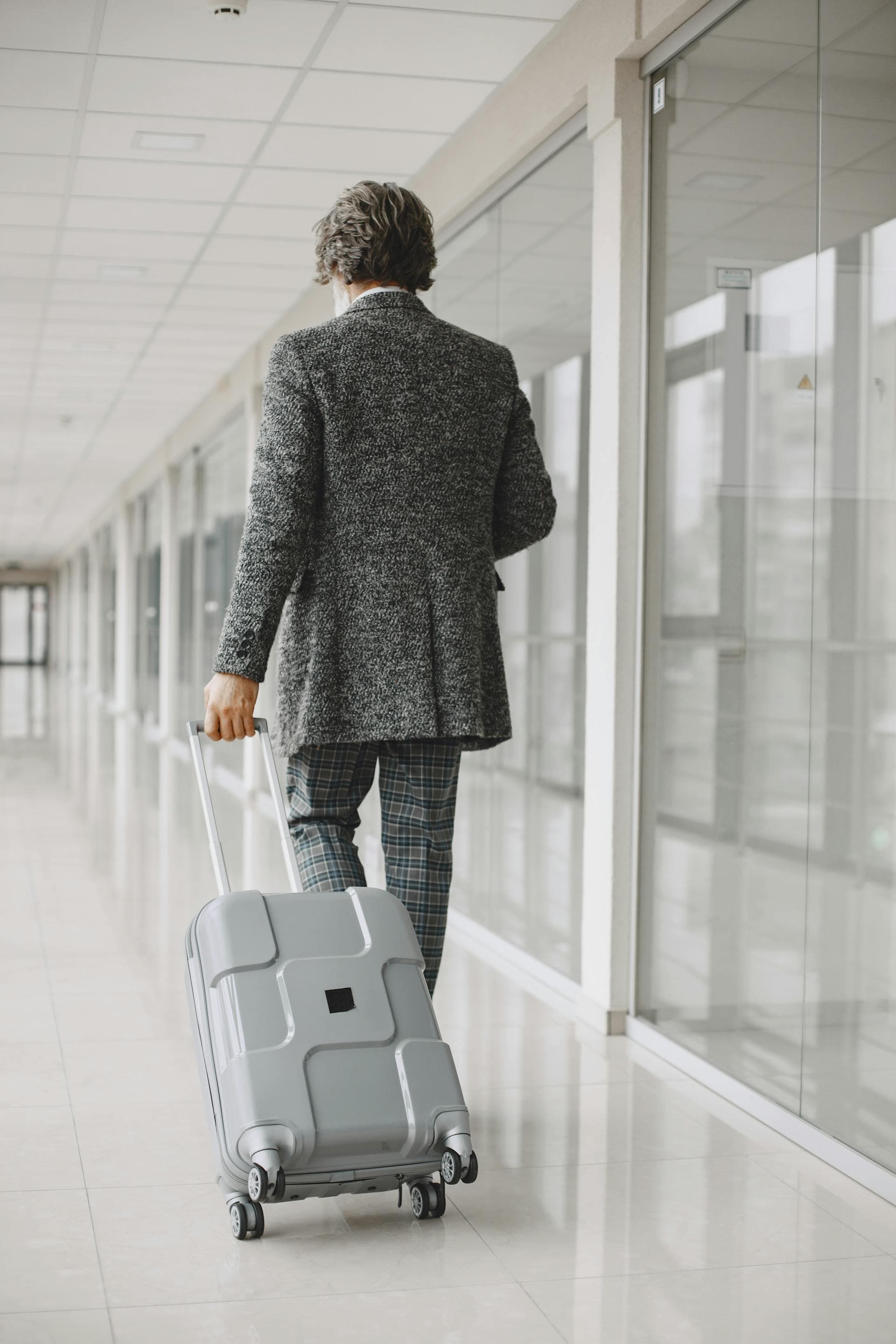 Businessman in a tailored coat walking through an airport corridor with rolling luggage