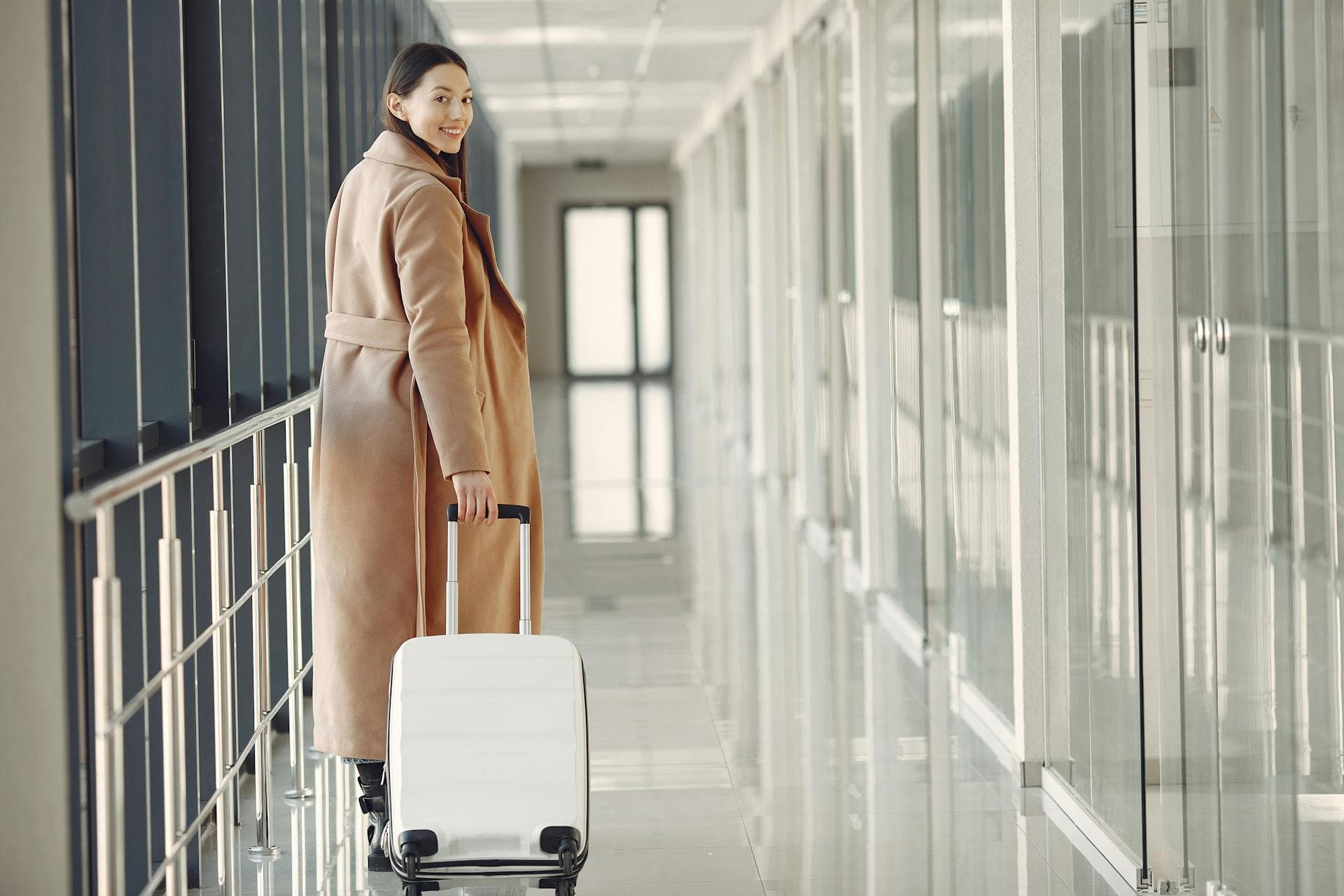 Solo traveler in a camel coat walking through a modern airport with a white carry-on